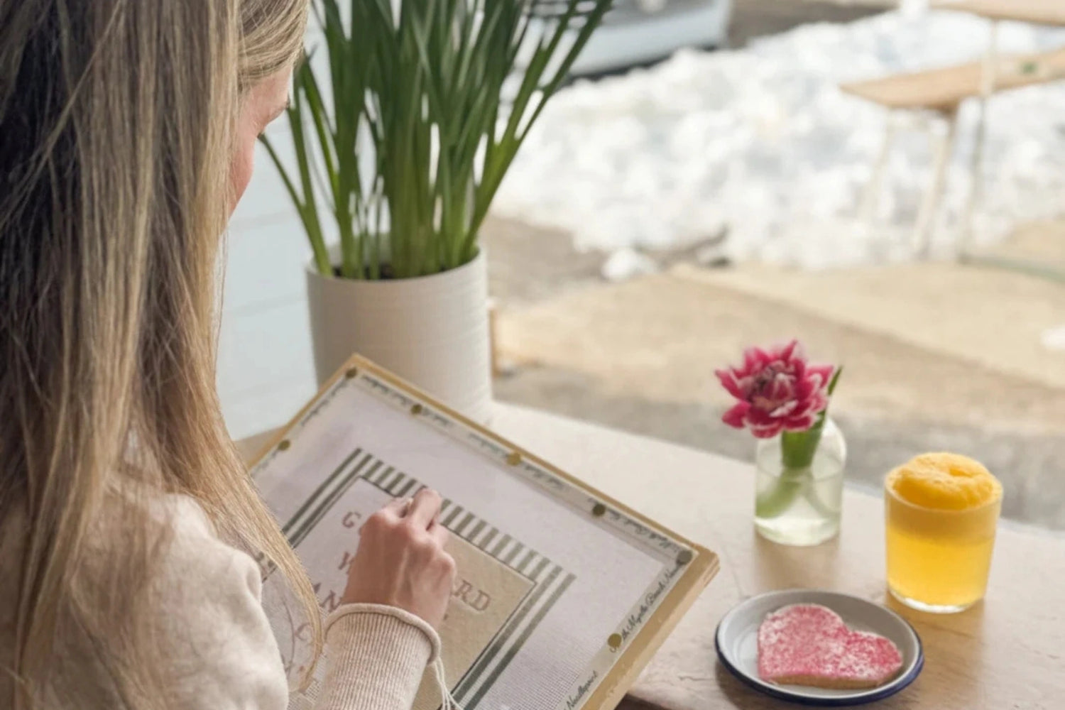 Woman stitching a needlepoint canvas at a cafe table with a drink and cookie.